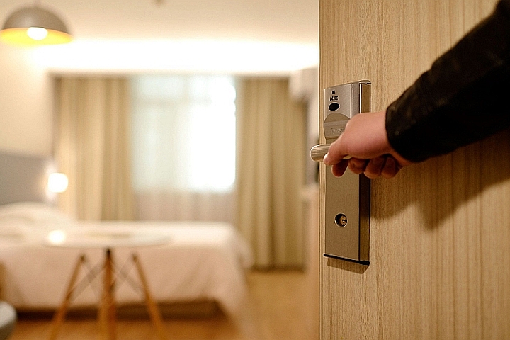Eine Hand öffnet eine Zimmertür, die in ein modernes Hotelzimmer führt. Im Hintergrund sind ein Bett und eine Fensterfront mit Vorhängen sichtbar. Der Raum wirkt hell und einladend.