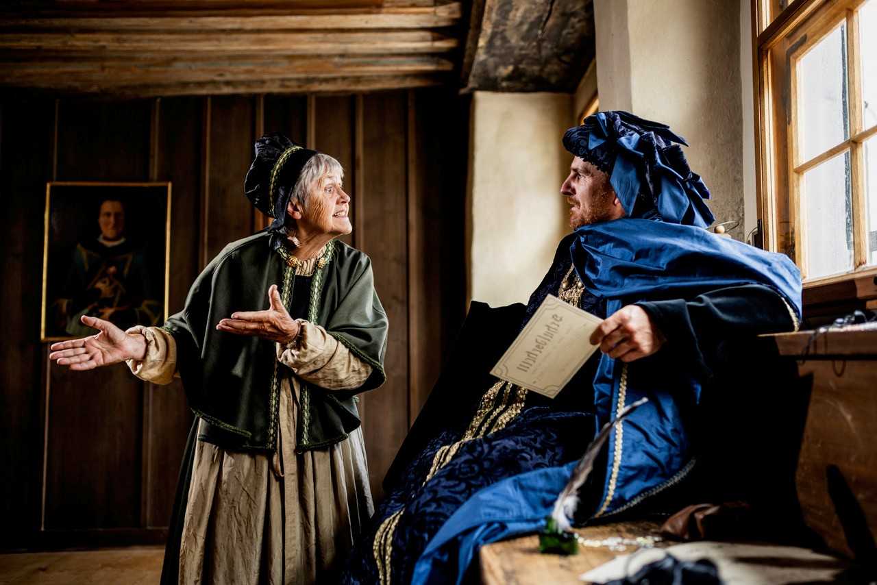 Zwei Personen in historischer Kleidung stehen in einem Raum mit Holzverkleidung. Eine Frau gestikuliert lebhaft, während ein Mann, der auf einem Tisch sitzt, ein Dokument in der Hand hält. Im Hintergrund ist ein Gemälde zu sehen.