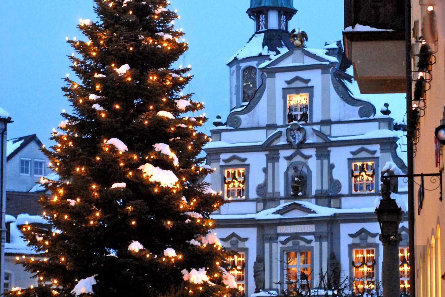 Ein weihnachtlich geschmückter Tannenbaum mit Lichtern und Schnee steht im Vordergrund. Im Hintergrund ist ein elegantes, historisches Gebäude zu sehen, das ebenfalls mit Schnee bedeckt ist und warme Lichter in den Fenstern leuchten. Der Himmel ist dunkelblau.