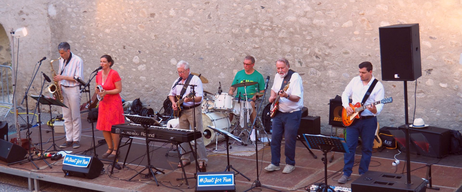 Eine Band spielt live auf einer Bühne im Freien. Sechs Musiker sind zu sehen, darunter ein Saxophonist, eine Sängerin in einem roten Kleid, ein Schlagzeuger und Gitarristen. Im Hintergrund sind Instrumente und Lautsprecher sichtbar.