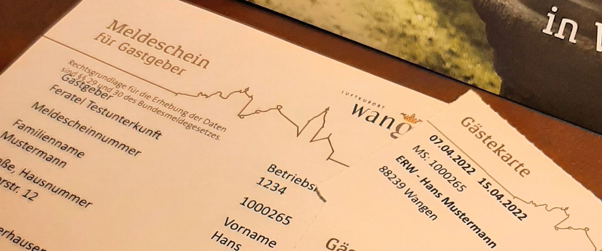 Eine Meldekarte für Gäste mit Feldern für die Unterkunft, Familienname, Vorname und Betriebsnummer. Die Karte enthält Platz für persönliche Daten, die angegeben werden müssen. Der Hintergrund zeigt eine natürliche Landschaft.