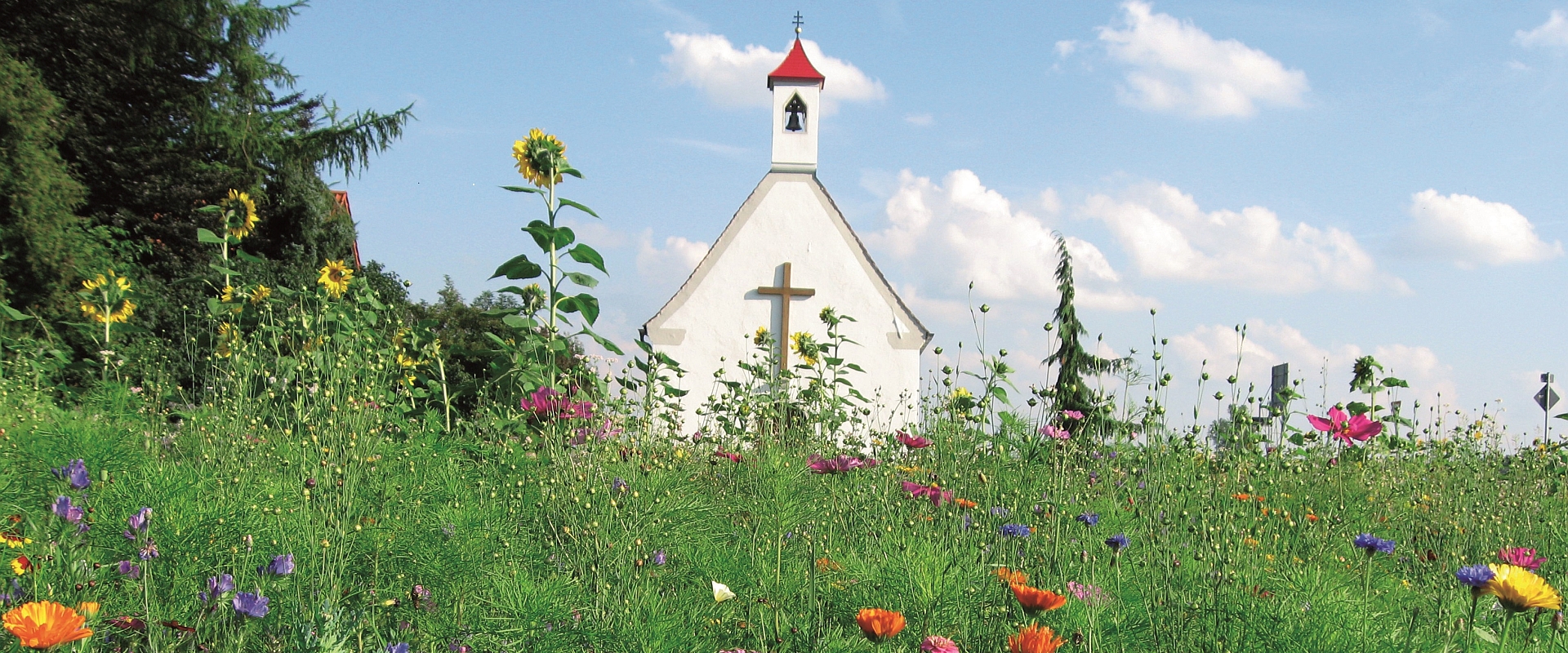 Eine weiße Kapelle mit rotem Dach und einem Kreuz steht im Hintergrund, umgeben von bunten Wildblumen und grünem Gras. Der Himmel ist blau mit einigen Wolken.