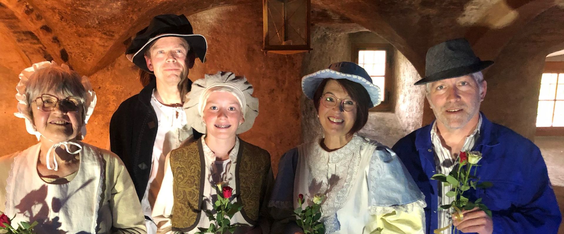 Fünf Personen in historischer Kleidung stehen in einem beleuchteten Raum. Sie halten jeweils eine Blume in der Hand und lächeln in die Kamera. Der Raum hat eine rustikale Atmosphäre mit sichtbaren Mauersteinen und einem warmen Lichtton.