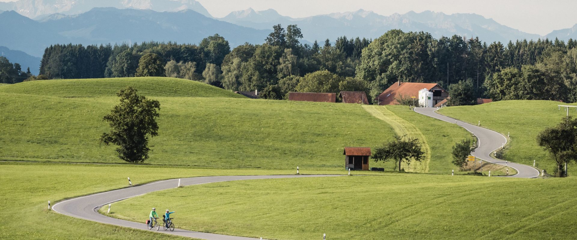 Zwei Radfahrer fahren auf einer kurvenreichen Straße durch grüne Wiesen, umgeben von Bäumen. Im Hintergrund sind sanfte Hügel und Berge sichtbar, unter einem klaren Himmel. Ein kleines Gebäude und eine Bushaltestelle stehen am Straßenrand.
