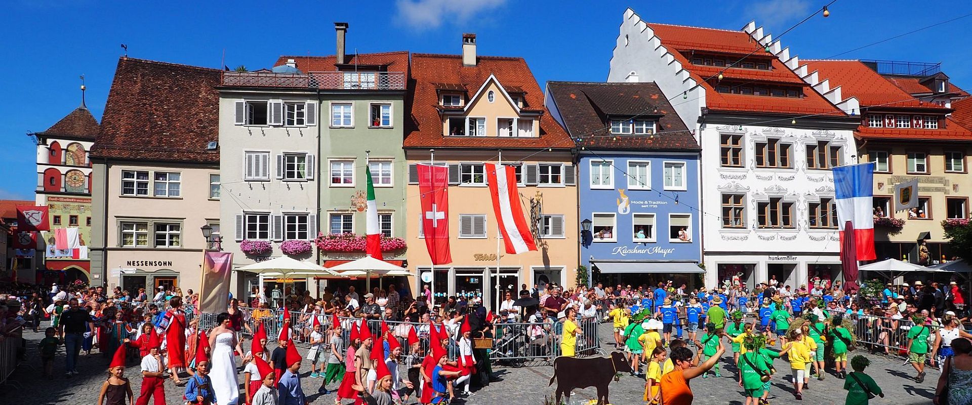 Eine belebte Stadtplatzszene mit bunten Festtagsumzügen. Menschen in farbenfrohen Kostümen ziehen zwischen historischen Gebäuden hindurch, während Zuschauer auf Terrassen sitzen. Fähnchen und Banner sind sichtbar, und die Atmosphäre ist festlich und lebhaft.