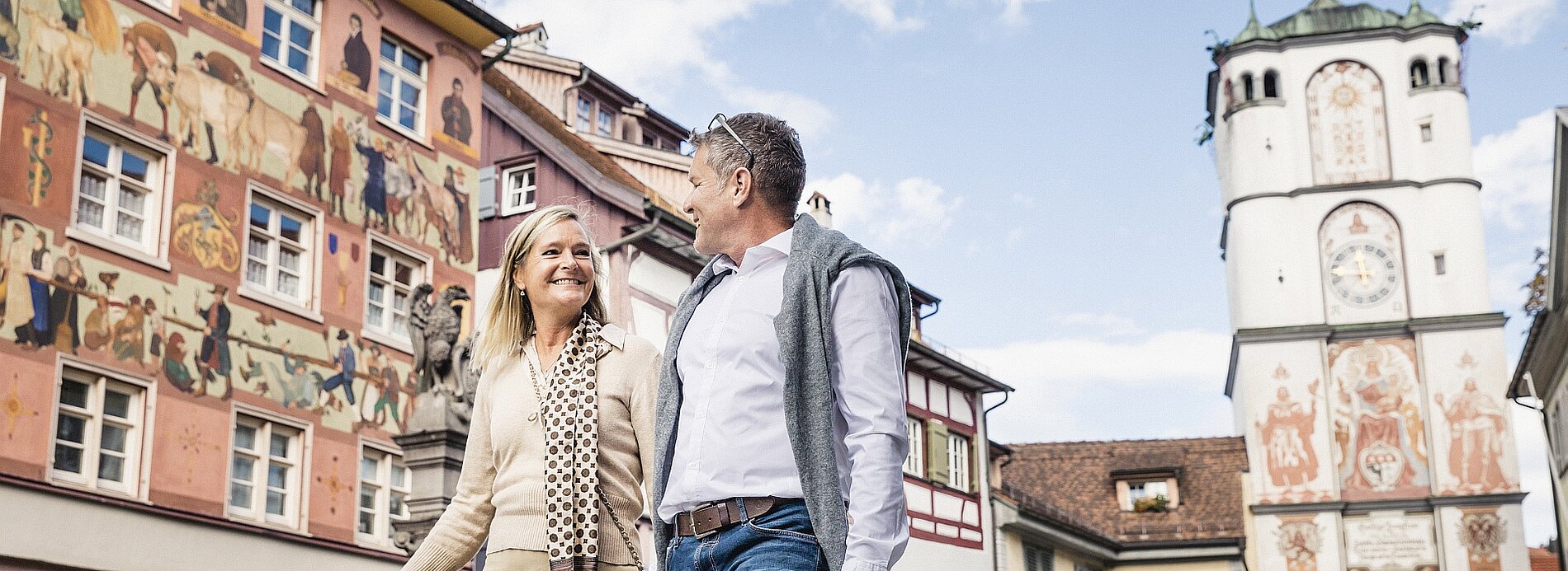 Ein Paar spaziert lächelnd durch eine charmante Altstadt mit historischen Gebäuden und Fresken. Im Hintergrund ist ein Uhrturm sichtbar, unter einem klaren blauen Himmel.
