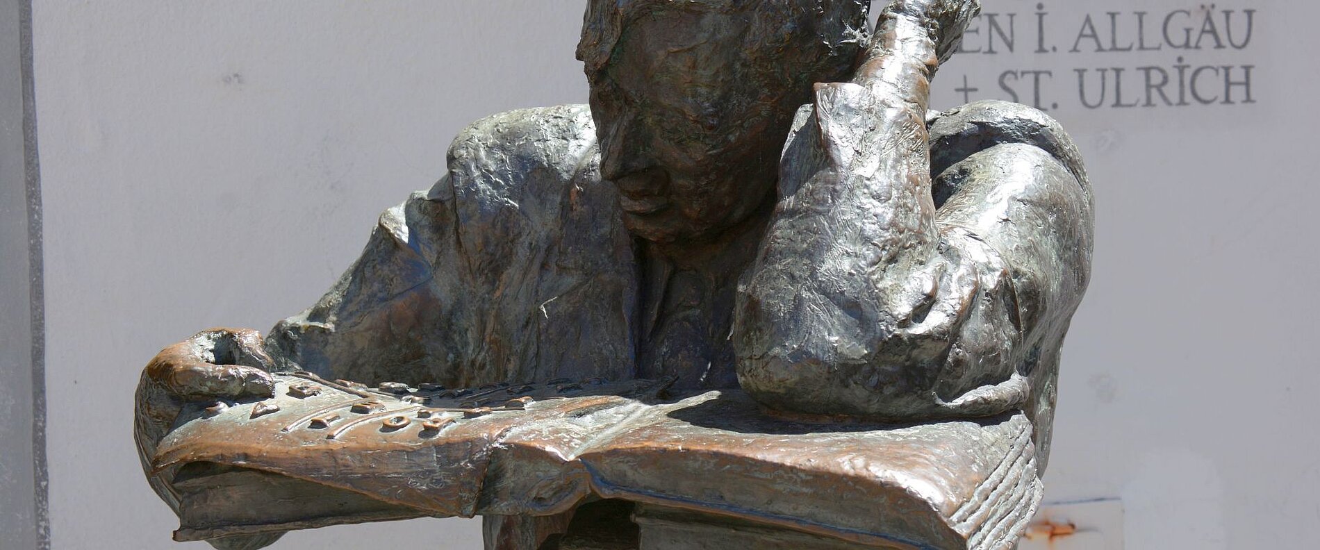 Eine bronzene Statue eines konzentrierten Mannes, der einen großen offenen Buch betrachtet und mit einer Hand an seinem Kopf stützt. Der Ausdruck zeigt Nachdenklichkeit. Im Hintergrund sind einige Schriftzüge sichtbar.