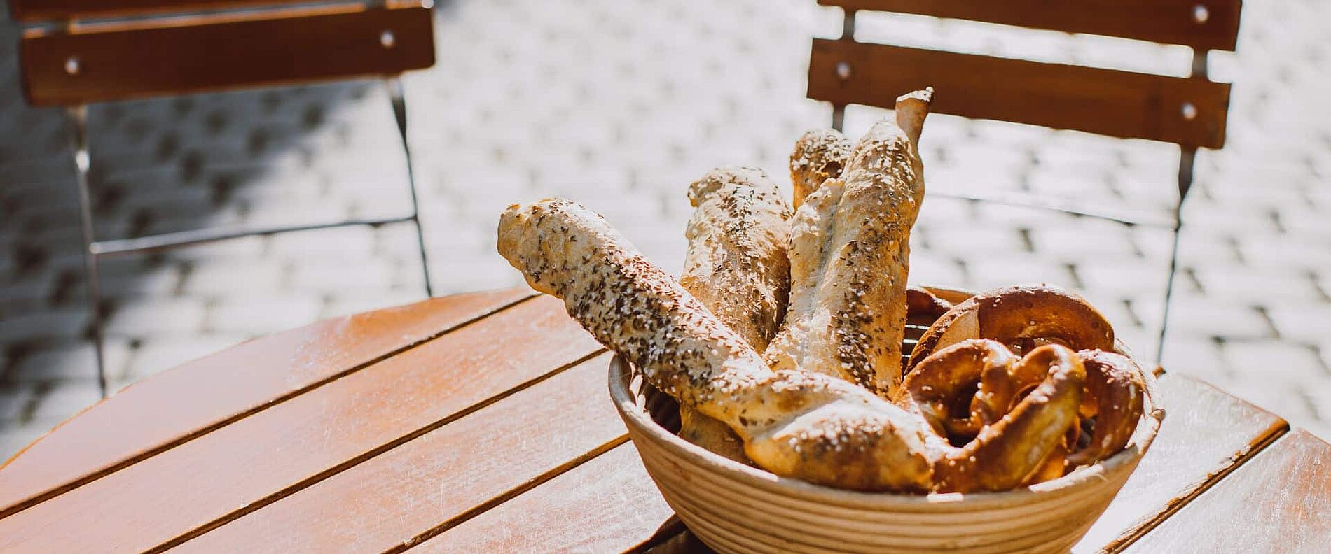 Ein Weidenkorb mit verschiedenen Brotstangen und Brezeln steht auf einem Holztisch. Im Hintergrund sind leere Stühle zu sehen und der Boden ist gepflastert. Das Bild zeigt eine einladende, sonnige Atmosphäre.