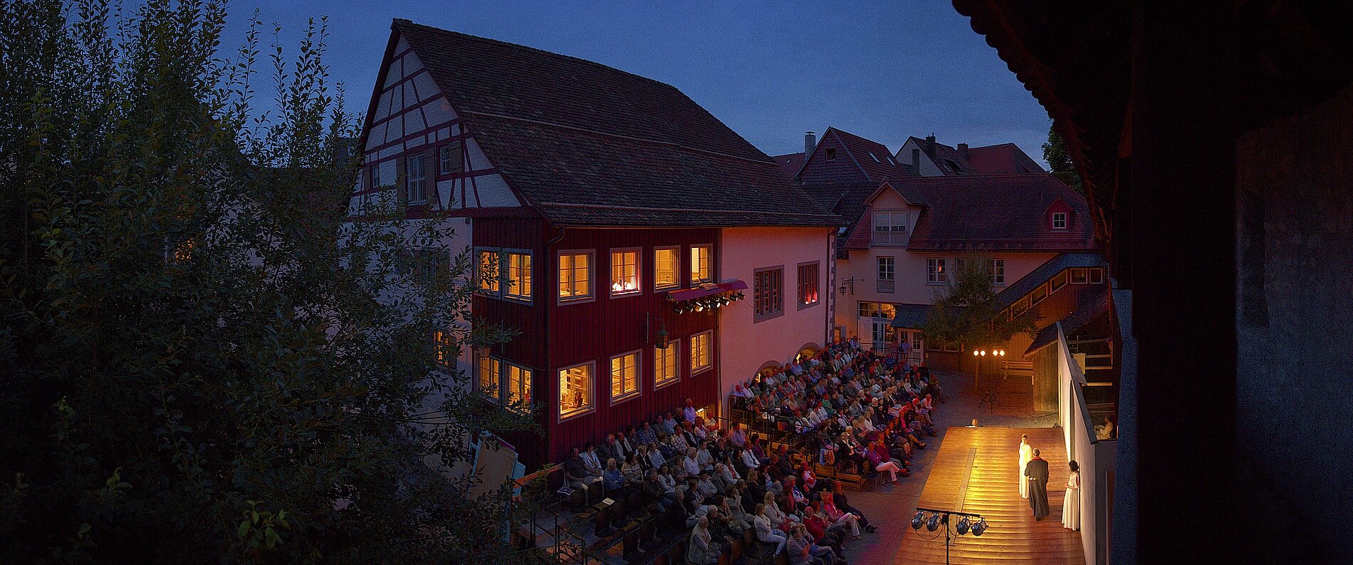 Eine belebte Freilichtbühne bei Dämmerung, umgeben von historischen Häusern. Zuschauer sitzen auf Bänken und schauen auf einen Redner, der auf der Bühne steht. Die Fenster der Häuser leuchten warm, während der Himmel langsam dunkel wird.