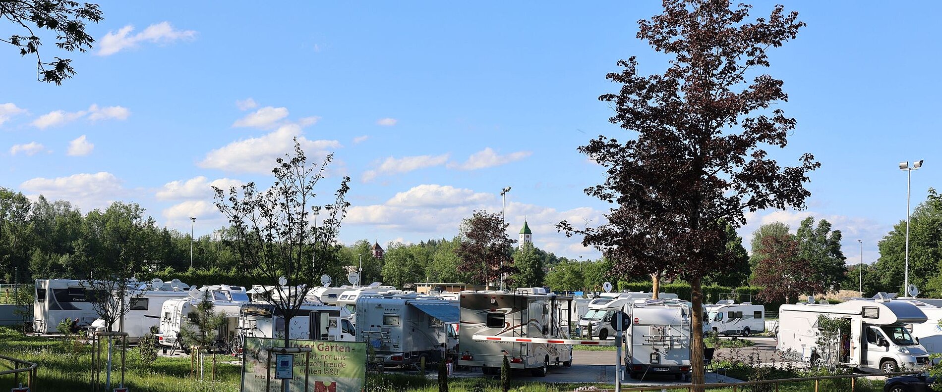 Ein Wohnmobilstellplatz mit mehreren Wohnmobilen, umgeben von Bäumen und einem blauen Himmel. Im Hintergrund sind einige Wolken sichtbar. Die Szene vermittelt eine ruhige, outdoor-orientierte Atmosphäre.