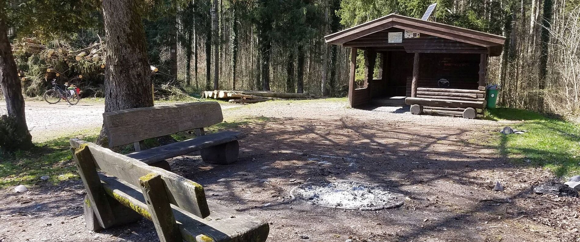 Ein rustikaler Unterstand im Wald mit Sitzbänken und einem leeren Feuerstelle. Im Hintergrund sind Bäume sichtbar und ein Radfahrer fährt vorbei. Die Umgebung ist sonnig und einladend.