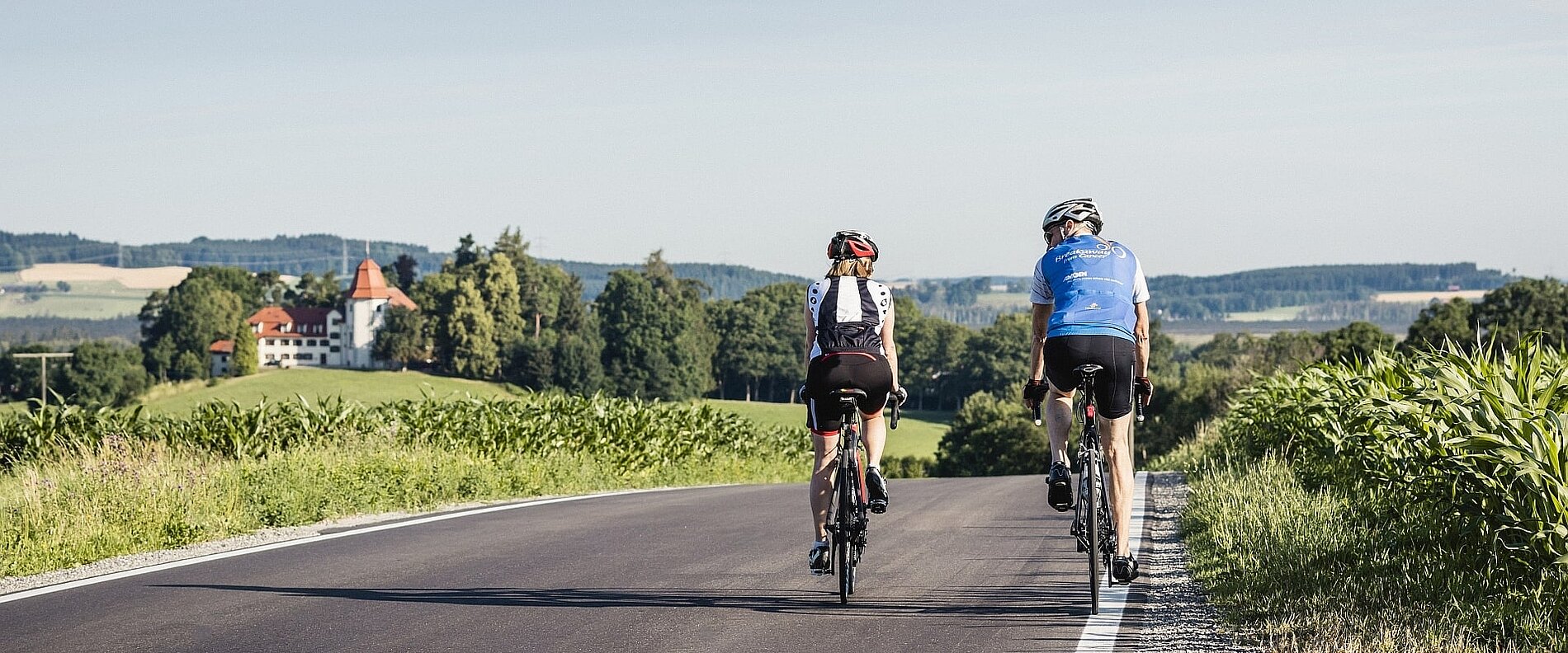Zwei Radfahrer fahren auf einer Landstraße, umgeben von grünem Feld und sanften Hügeln. Im Hintergrund ist ein schönes Bauernhaus mit rotem Dach sichtbar. Die Landschaft ist sonnig und friedlich.