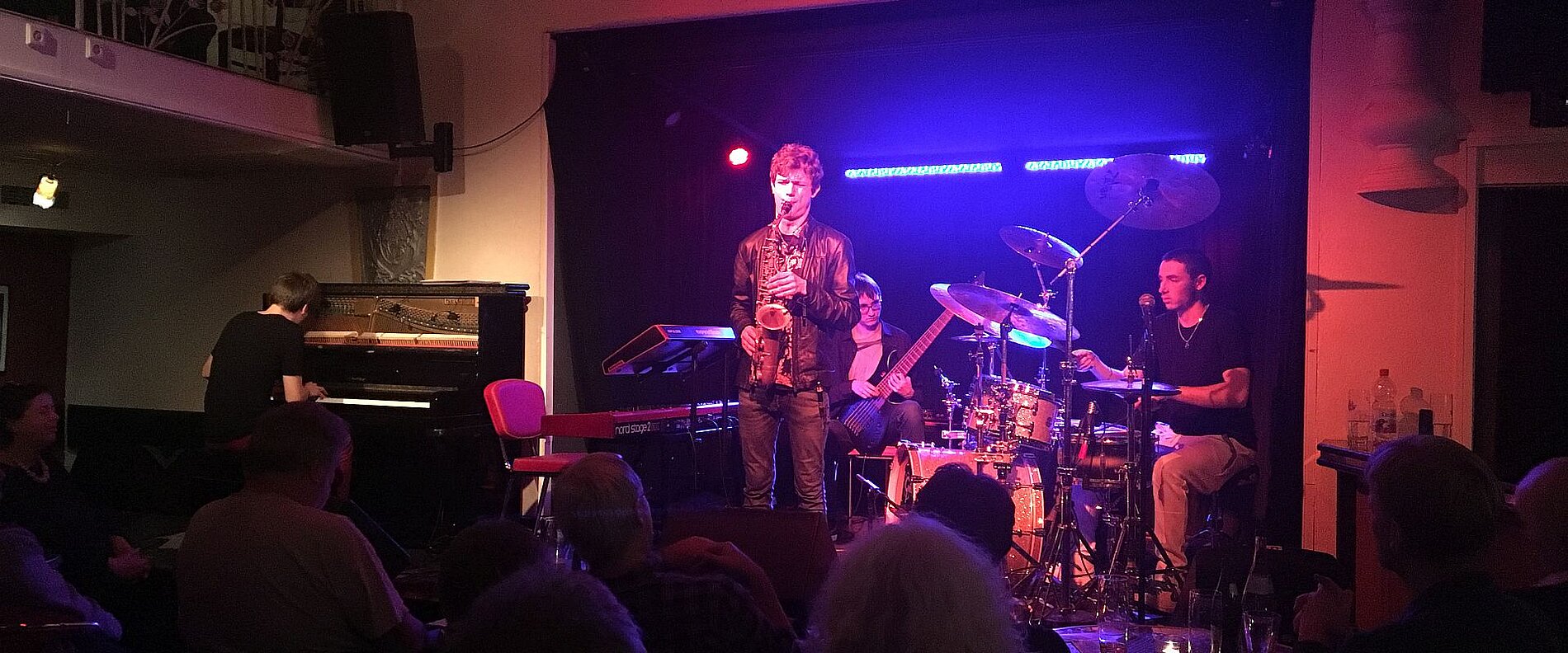 Eine Band spielt live auf einer Bühne in einem Nachtclub. Ein Sänger steht im Vordergrund, während Musiker an einem Klavier und Schlagzeug performen. Das Publikum sitzt in der Nähe und hört gespannt zu. Die Bühne ist in buntem Licht beleuchtet.