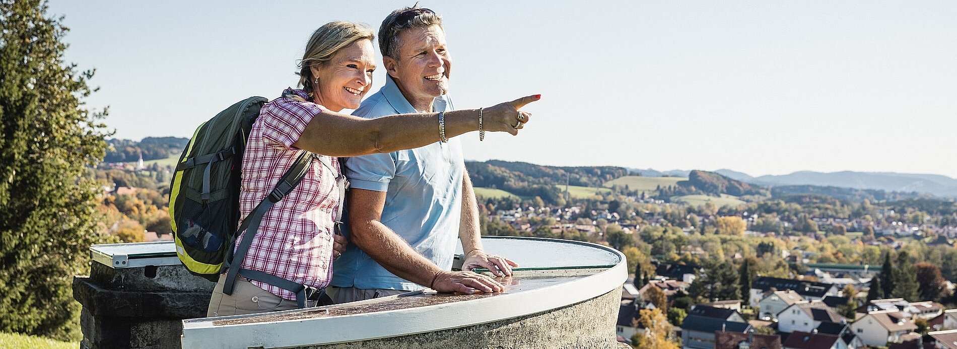 Ein Paar steht an einem Aussichtspunkt und blickt auf die Landschaft. Die Frau zeigt mit dem Finger auf etwas in der Ferne, während der Mann lächelt. Im Hintergrund sind sanfte Hügel und ein Dorf sichtbar. Es ist ein sonniger Tag.