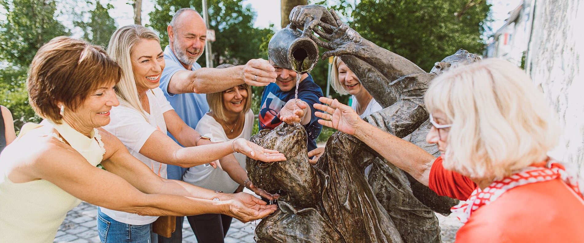 Eine Gruppe von sieben Menschen umkreist eine Statue und hält die Hände unter einen Wasserkrug, von dem Wasser fließt. Sie lächeln und scheinen eine interaktive Aktivität zu genießen, während eine sonnige Umgebung im Hintergrund zu sehen ist.