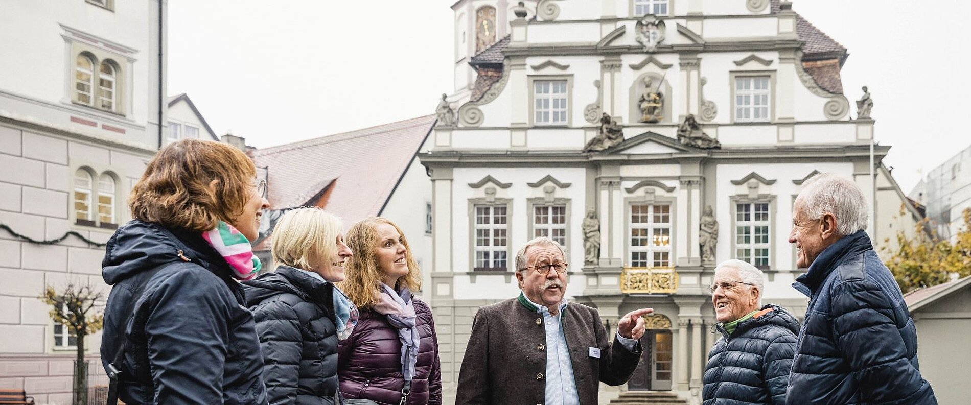 Sechs Personen stehen in einer Stadtansicht. Eine Person erklärt etwas, während die anderen gebannt zuhören. Im Hintergrund ist ein historisches Gebäude mit dekorativen Elementen zu sehen. Der Himmel ist bewölkt und die Umgebung wirkt herbstlich.