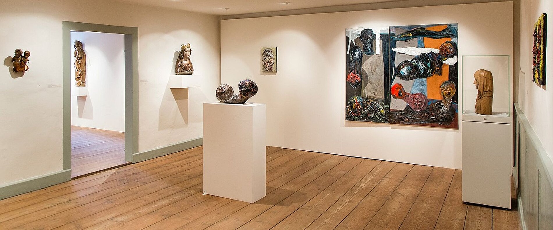 Ein Innenraum einer Galerie mit Holzfußboden. An den Wänden hängen verschiedene Kunstwerke, darunter Gemälde und Skulpturen. In der Mitte des Raums steht ein weißer Sockel mit einer weiteren Skulptur. Der Raum wirkt hell und einladend.