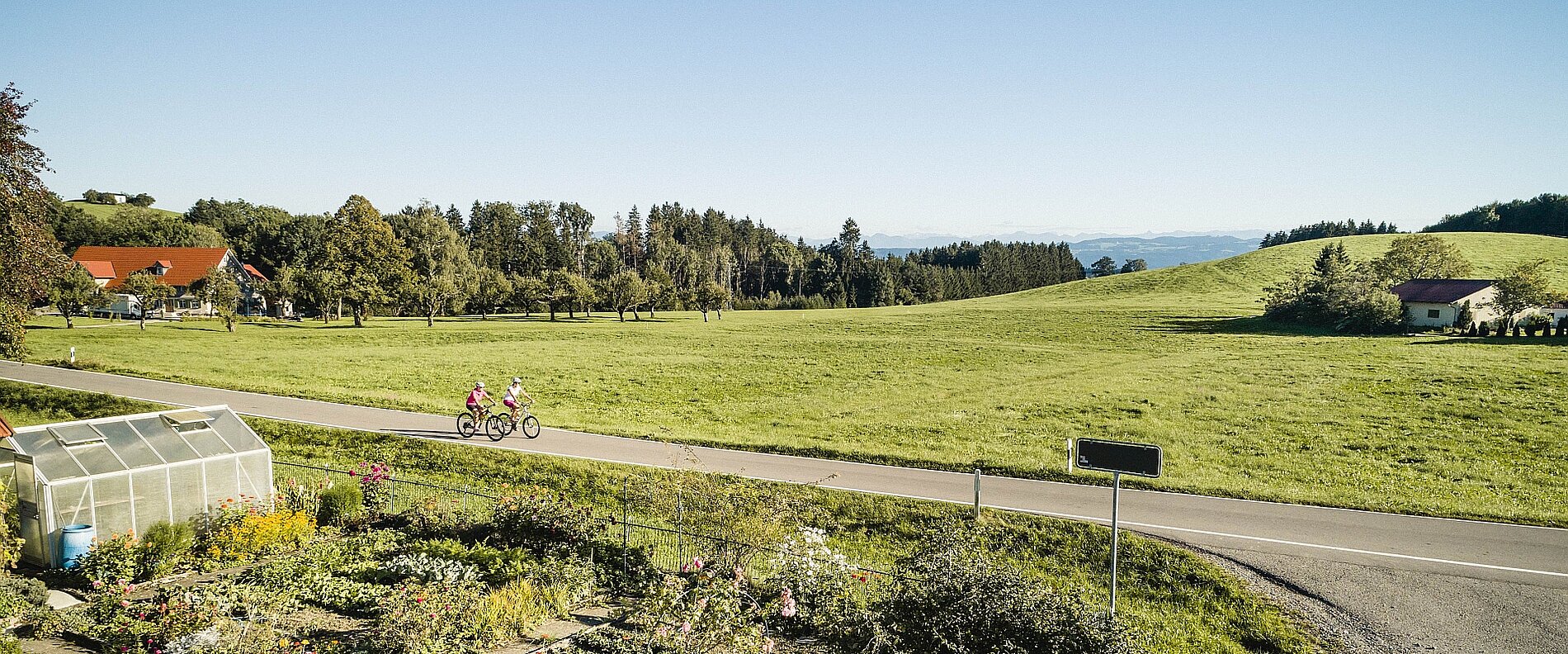 Zweiradfahrer fahren eine Straße entlang, umgeben von weiten grünen Wiesen und Bäumen. Im Vordergrund ist ein Garten mit Blumen und einem Gewächshaus zu sehen. Im Hintergrund erheben sich sanfte Hügel und eine klare blaue Himmel.