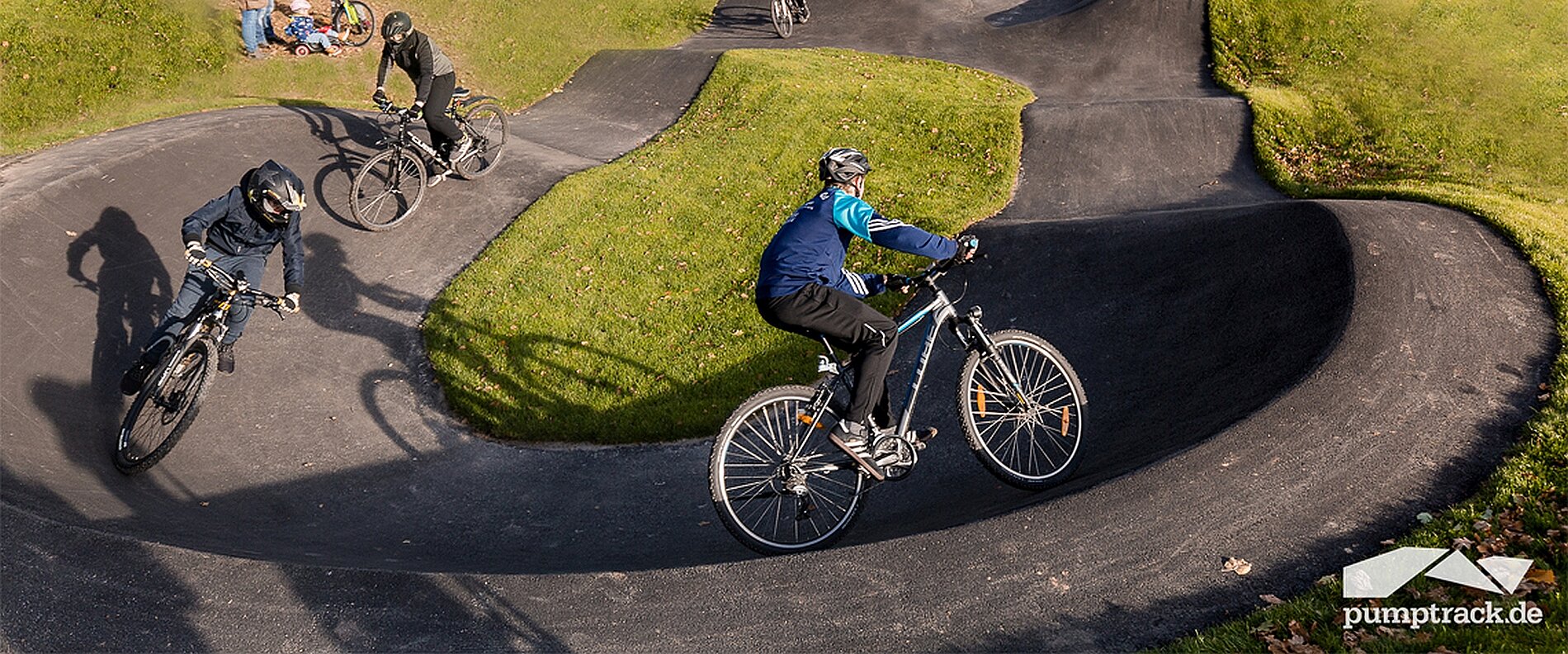 Mehrere Radfahrer fahren auf einem Pumptrack mit geschwungenen, grünen Plattformen. Die Fahrer tragen Helme und Radbekleidung. Im Hintergrund sind Zuschauer auf einer Wiese zu sehen. Der Boden ist asphaltiert und die Umgebung ist grün.