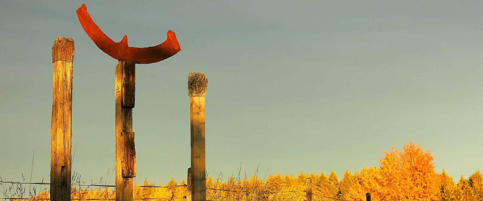Ein rostiges gebogenes Metallstück ist auf einem Holzpfahl montiert. Daneben stehen drei weitere Holzpfähle, während im Hintergrund ein goldenes Herbstlicht die Landschaft erhellt. Bäume in herbstlichen Farben sind sichtbar.