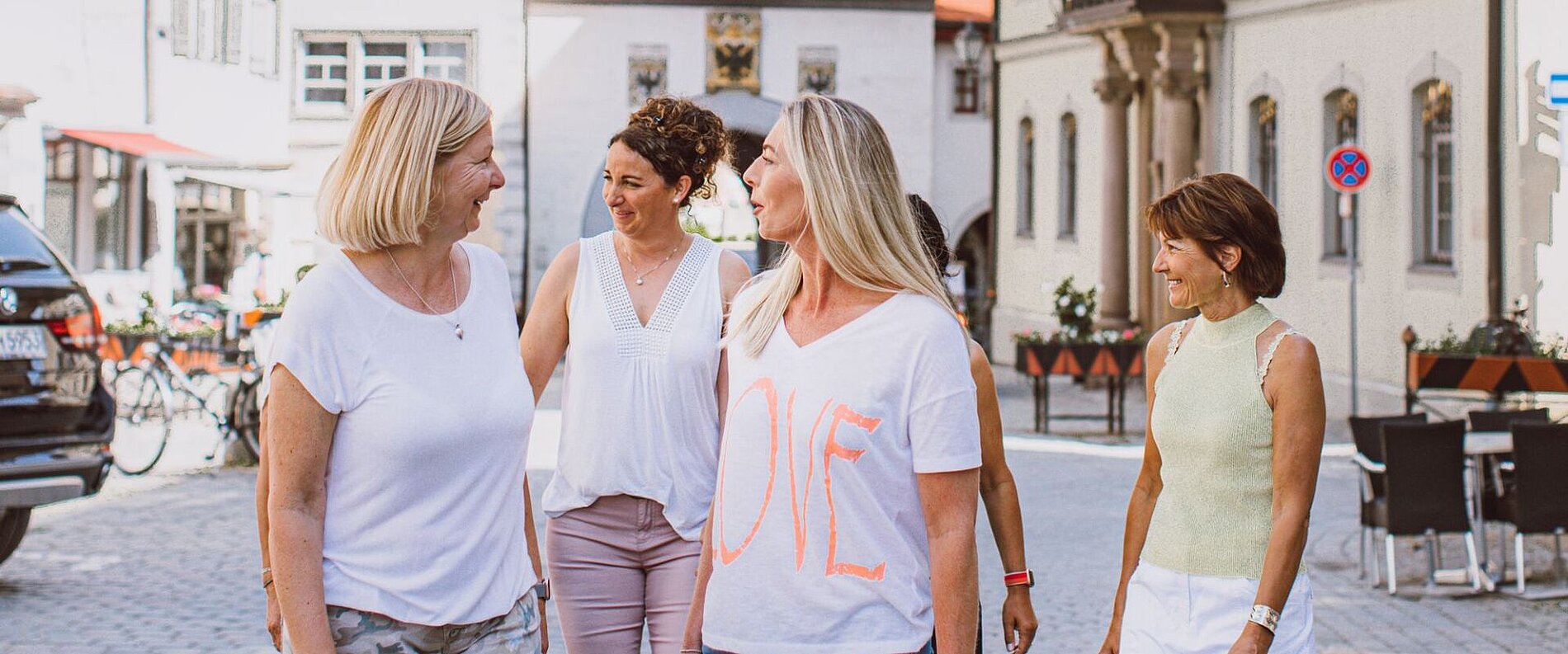 Fünf Frauen spazieren in einer Stadtstraße. Sie tragen unterschiedliche, sommerliche Kleidung und lächeln einander an. Im Hintergrund sind Gebäude und parkende Autos zu sehen.