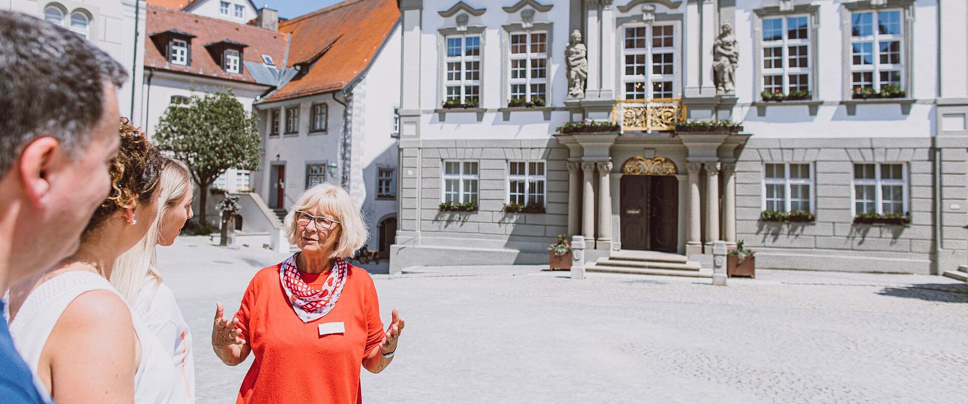 Eine Gruppe von vier Personen hört aufmerksam einer älteren Frau in rotem Oberteil zu. Sie steht vor einem historischen Gebäude mit Skulpturen und einem Balkon. Die Szenerie spielt sich auf einem gepflasterten Platz ab, an einem sonnigen Tag.