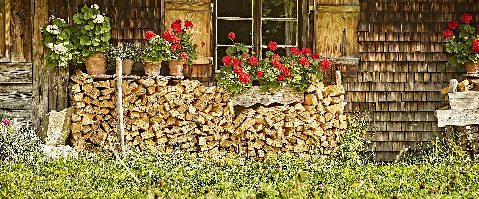 Ein holzverkleideter Außenbereich eines Hauses mit gestapeltem Brennholz. Darauf stehen Töpfe mit roten Blumen. Die Wände sind aus Holz und das Fenster ist sichtbar, mit Holzläden an den Seiten. Im Vordergrund wächst grünes Gras.