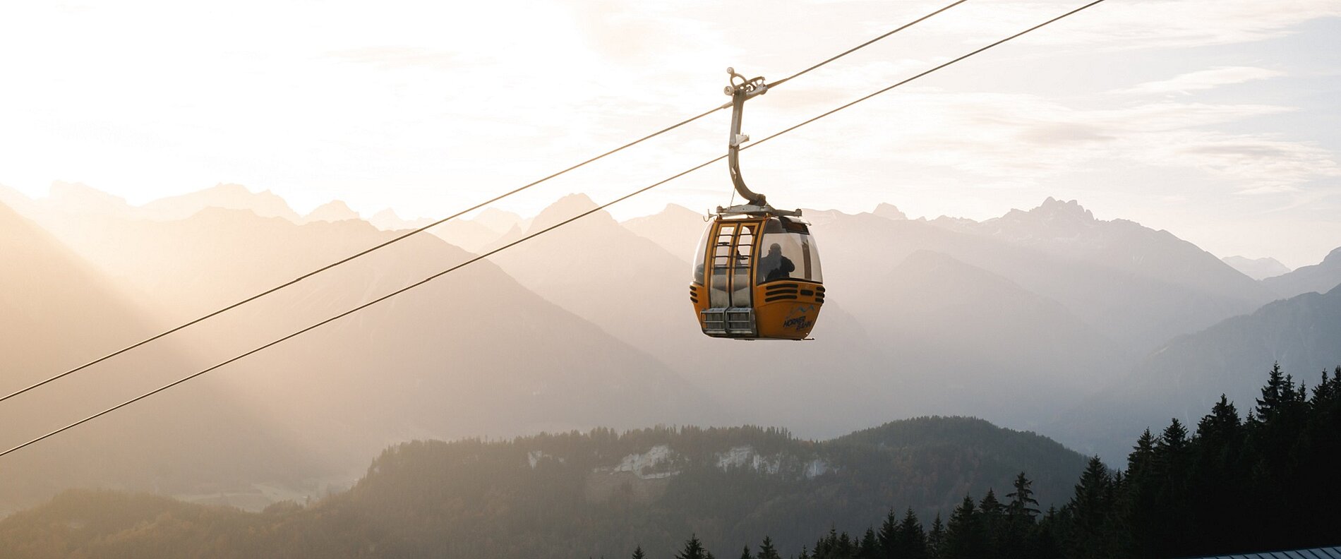 Eine Seilbahn schwebt über eine bergige Landschaft, während die Sonne hinter den Bergen aufgeht. Der Himmel hat einen sanften Farbverlauf und die Silhouetten der Berge sind im Hintergrund sichtbar. Die Umgebung ist von Bäumen umgeben.