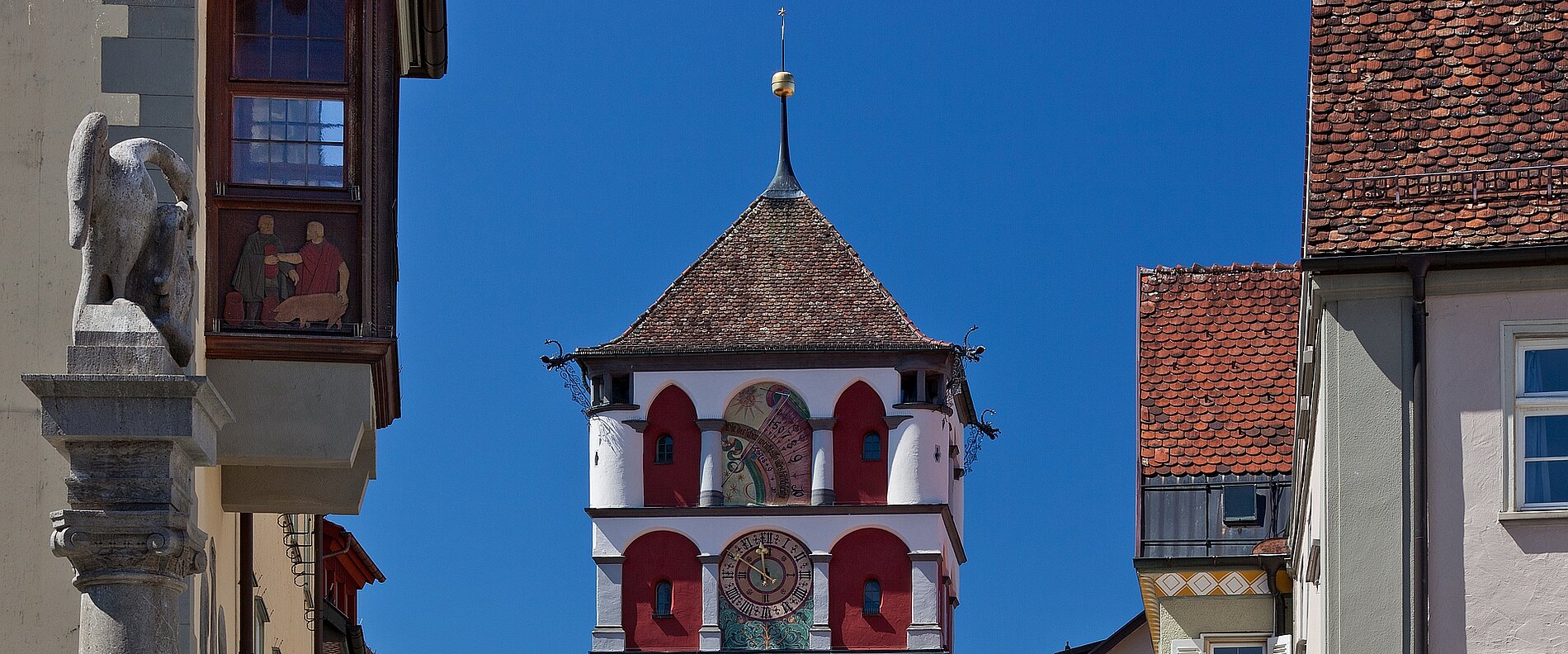 Blick auf einen historischen Stadtturm mit einem rot-weißen Design unter klarem blauem Himmel, umgeben von alten Gebäuden und einem Steinlöwen auf der linken Seite.