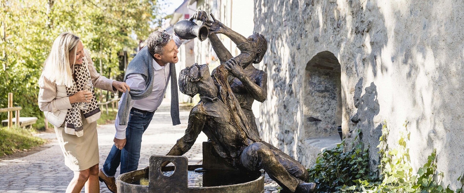 Eine Frau und ein Mann stehen vor einer Statue, die einen Mann darstellt, der aus einem Krug Wasser in einen Eimer gießt. Die Frau wirkt interessiert, während der Mann nahe an die Statue tritt. Im Hintergrund sind Bäume und eine Mauer sichtbar.