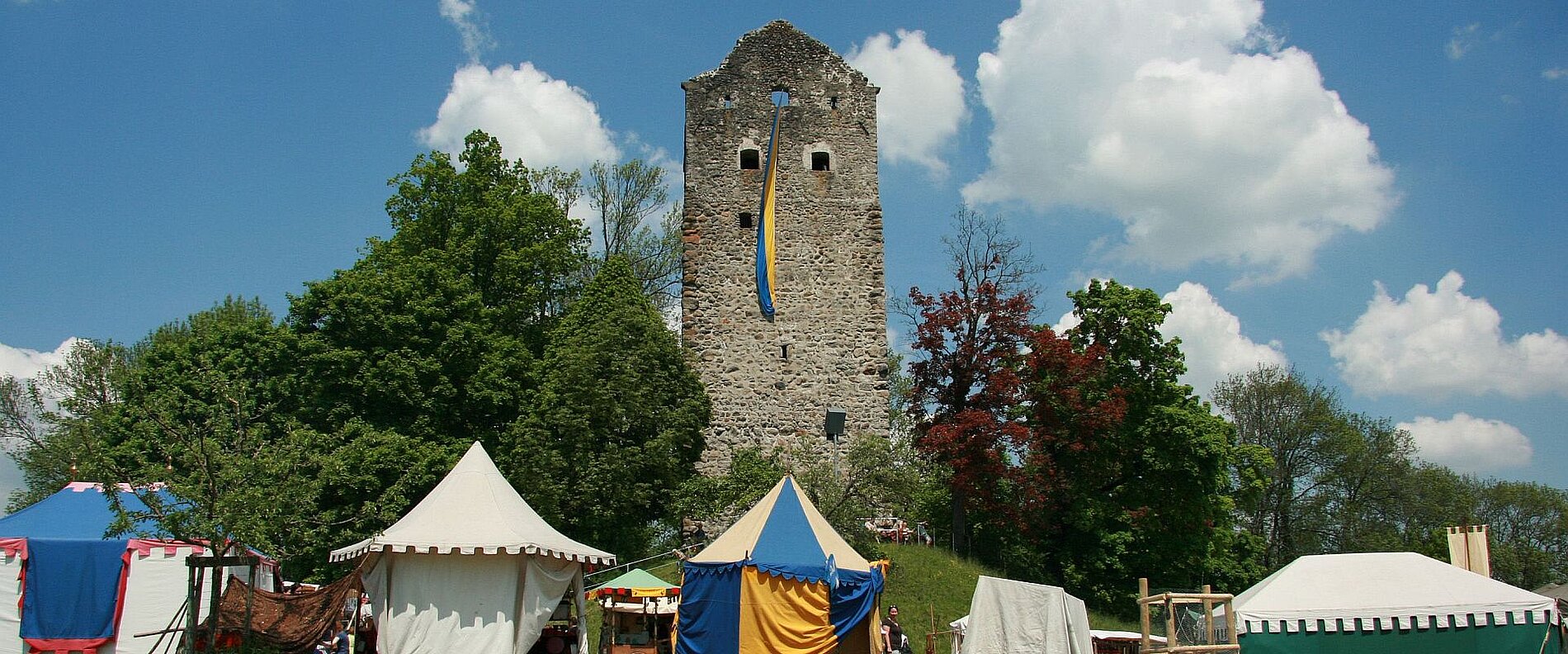 Eine alte Burgruine steht vor einem blauen Himmel mit Wolken. Im Vordergrund sind mehrere bunte Zelte mit verschiedenen Formen und Farben zu sehen, umgeben von Bäumen. Der Anblick vermittelt eine medievial anmutende Atmosphäre.