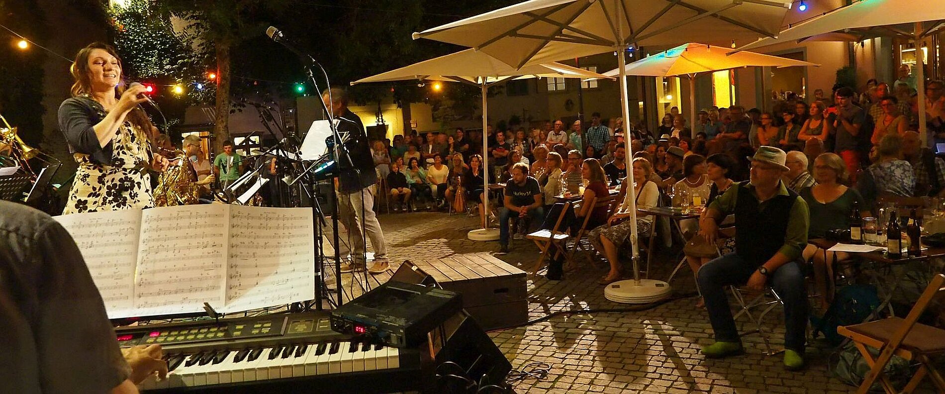 Eine Musikveranstaltung im Freien, bei der eine Künstlerin mit Mikrofon auftritt. Im Hintergrund ist ein Publikum zu sehen, das auf Stühlen sitzt und den darbietenden Künstlern zuhört. Die Umgebung ist festlich beleuchtet mit Sonnenschirmen und bunten Lichtern.