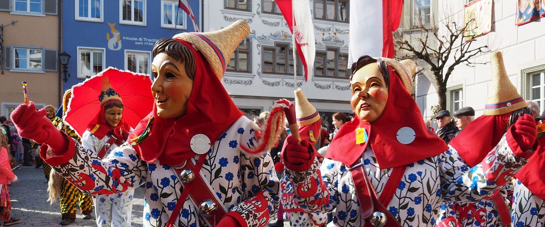 Bunt verkleidete Menschen mit großen Masken und Hüten nehmen an einem Festumzug teil. Einige Teilnehmer halten Schirme und andere Requisiten, während sie fröhlich tanzen und feiern. Hintergrund zeigt historische Gebäude und Zuschauer.