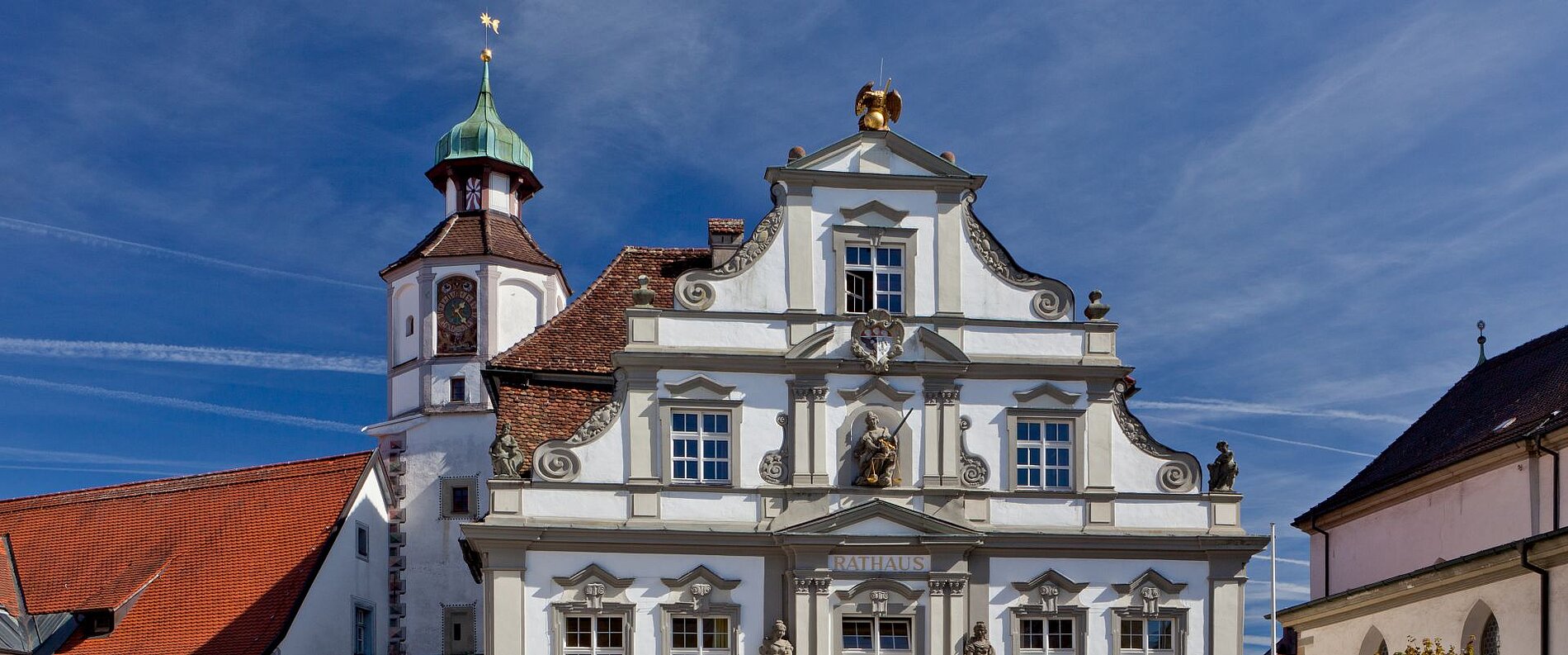 Ein historisches Gebäude mit einer eleganten, symmetrischen Fassade, geschmückt mit Statuen und goldenen Verzierungen. Oben steht ein kleiner Turm mit einer Kuppel. Der Himmel ist blau mit einigen Wolkenlinien.