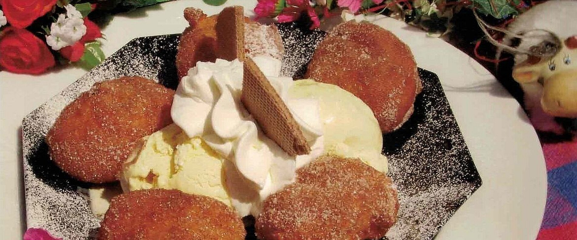 Ein schwarz-weißer Teller mit frittierten Süßigkeiten, umgeben von Vanilleeis und dekoriert mit Schlagsahne und einer Waffel. Im Hintergrund sind Blumen zu sehen.