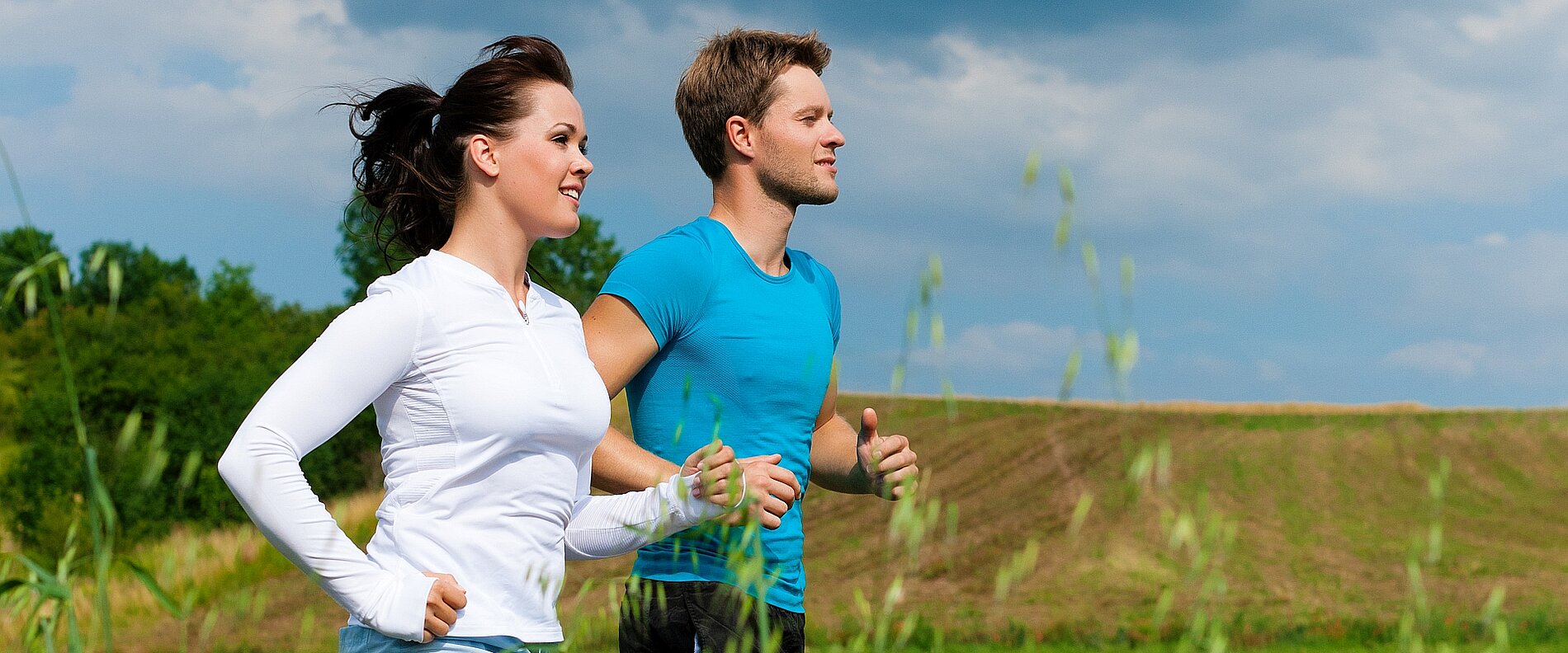 Ein Mann und eine Frau joggen gemeinsam im Freien. Beide tragen Sportkleidung und lächeln, während sie an einer grünen Wiese vorbeilaufen. Der Himmel ist teilweise bewölkt.