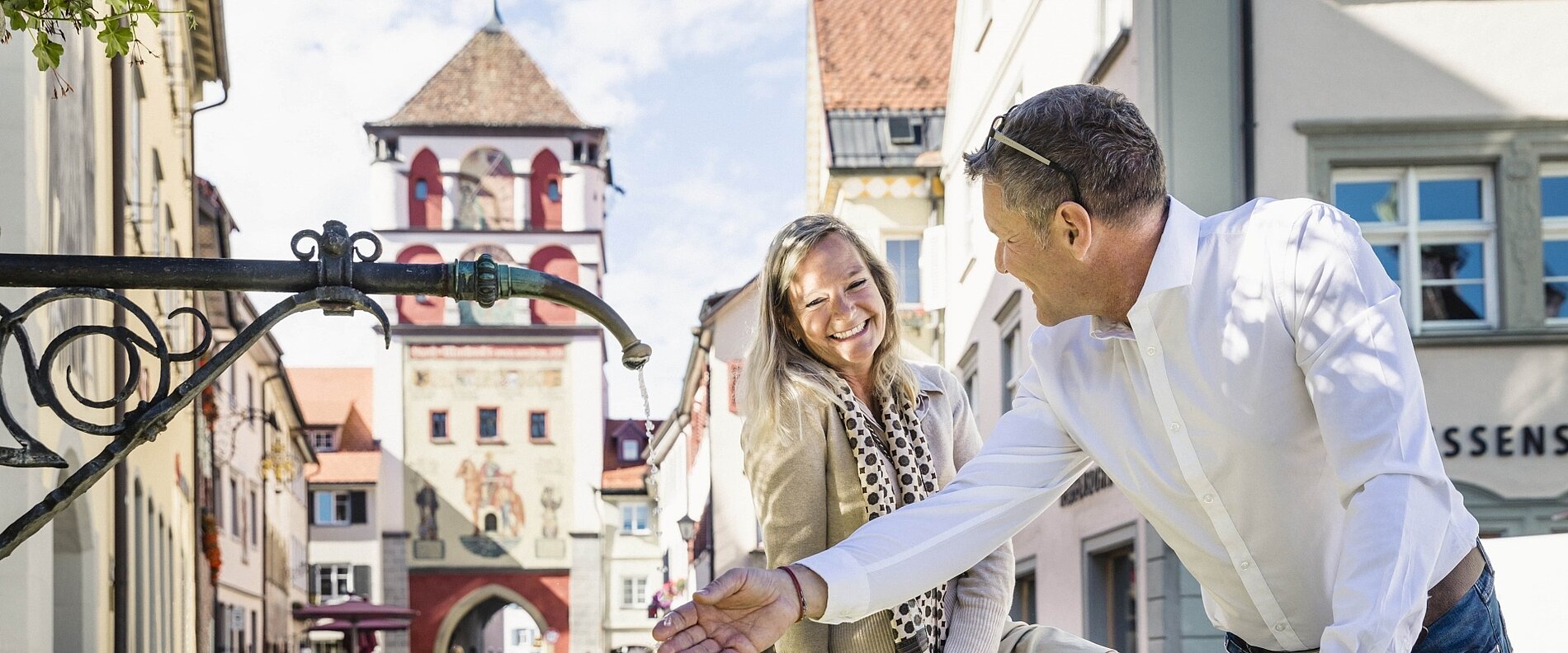 Zwei Personen stehen in einer engen Straße mit historischen Gebäuden im Hintergrund. Sie lächeln und scheinen einander freundlich zu begegnen, während ein Turm mit einem roten Ziffernblatt sichtbar ist. Die Atmosphäre wirkt einladend und gemütlich.