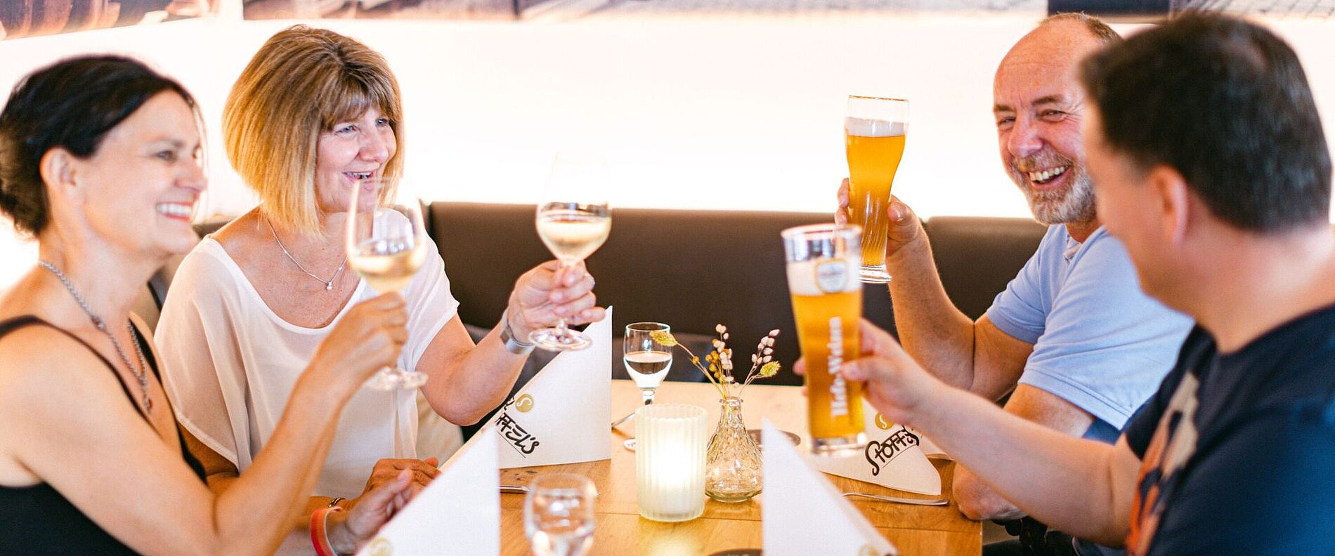 Vier Personen sitzen an einem Tisch in einem Restaurant und stoßen mit Getränken an. Zwei Frauen halten Weingläser, während ein Mann und eine Frau Biergläser heben. Die Atmosphäre ist freundlich und feierlich.