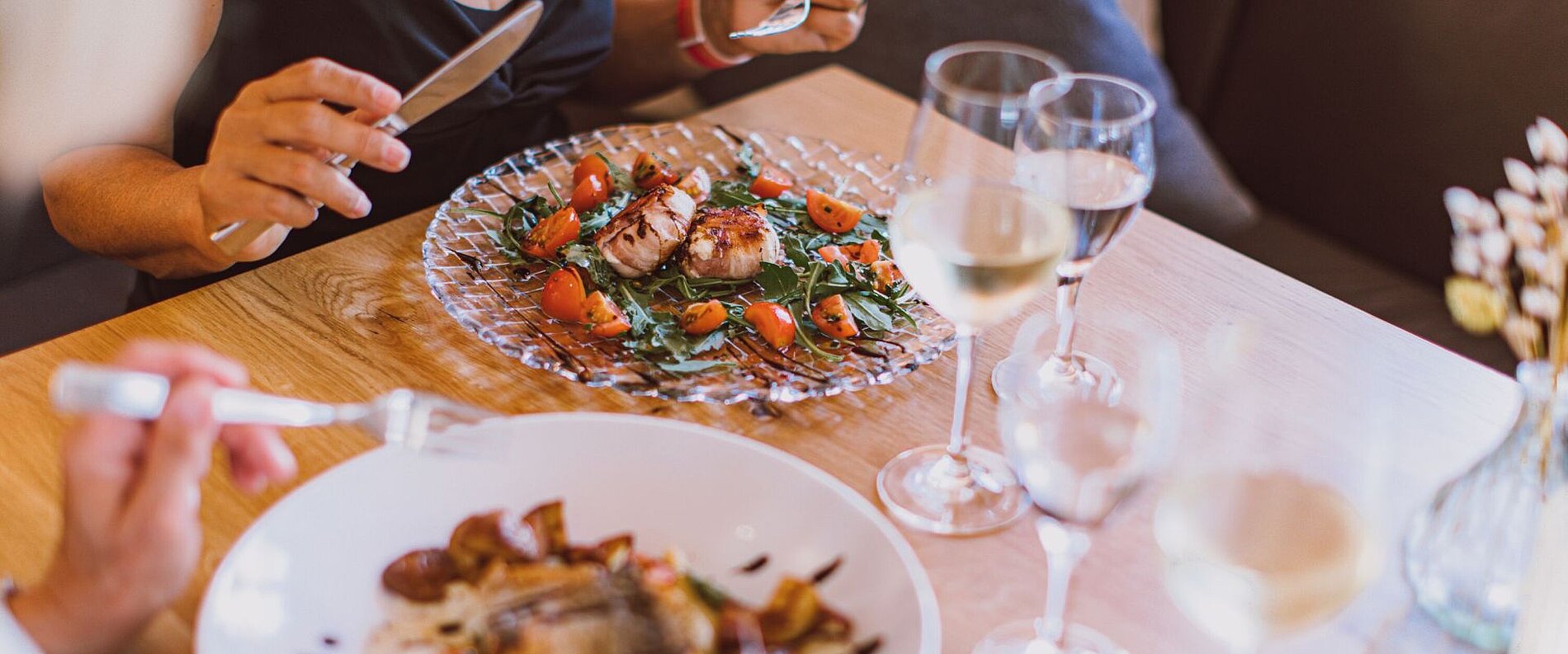 Ein Tisch mit zwei Tellern Essen, einem gegrillten Fischgericht und einem Salat mit Tomaten. Es stehen Gläser mit Weißwein auf dem Tisch. Eine Person hält eine Gabel und eine andere Person hat ein Messer in der Hand.