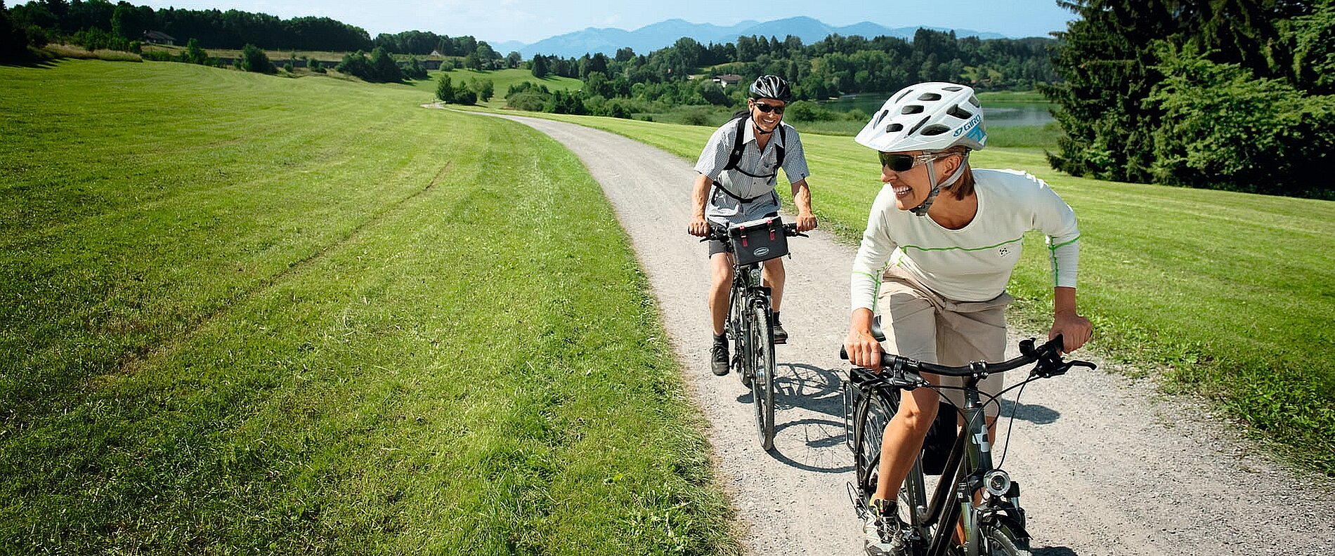 Zwei Männer radeln auf einem geschotterten Weg durch eine grüne Landschaft. Im Hintergrund sind sanfte Hügel und ein See sichtbar. Der Mann vorne trägt einen Fahrradhelm und lächelt, während der andere behindert ist.