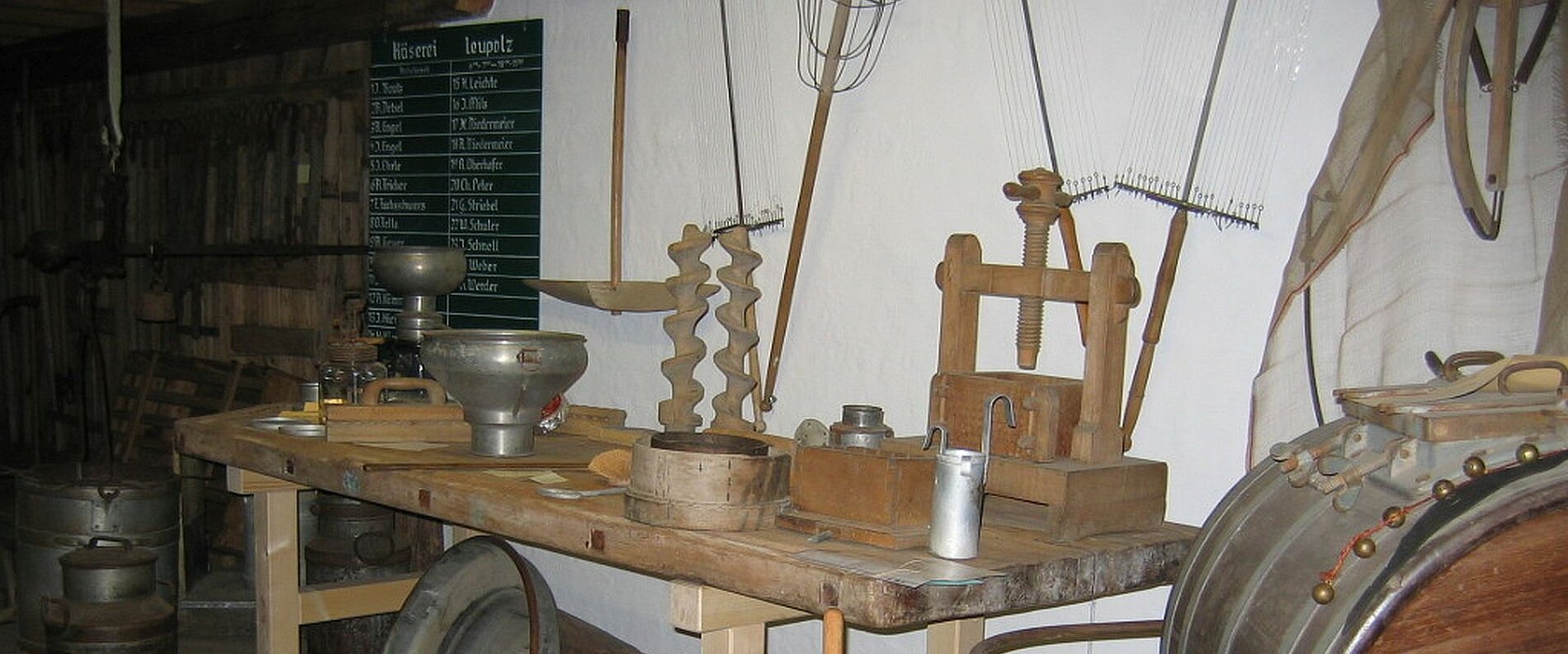Auf einem Tisch stehen verschiedene traditionelle Handwerkszeuge, darunter ein Trichter, eine Spirale, eine Holzpresse und einige Metallbehälter. Im Hintergrund ist eine grüne Tafel mit Informationen zu finden. Die Umgebung wirkt rustikal und erinnert an alte Werkstattpraktiken.
