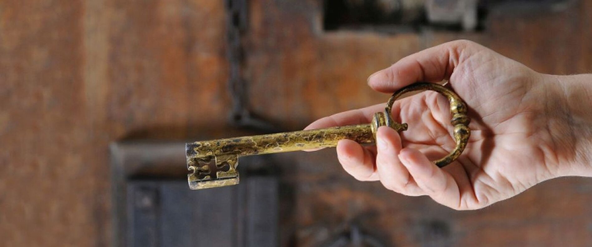 Eine Hand hält einen großen, goldenen Schlüssel vor einer rustikalen Hintergrundoberfläche. Der Schlüssel hat eine dekorative Form und scheint alt zu sein.