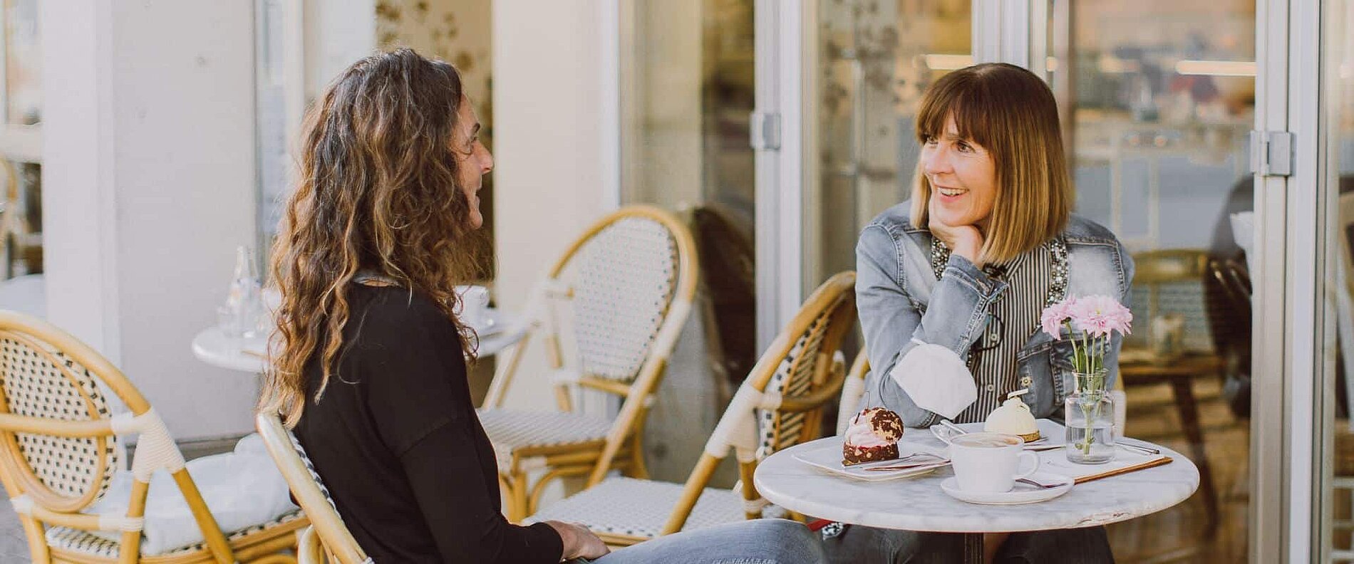 Zwei Frauen sitzen an einem kleinen Tisch in einem Café. Sie lächeln sich an und unterhalten sich. Auf dem Tisch steht eine Tasse Kaffee und ein Stück Kuchen. Im Hintergrund sind Stühle und Pflanzen sichtbar.