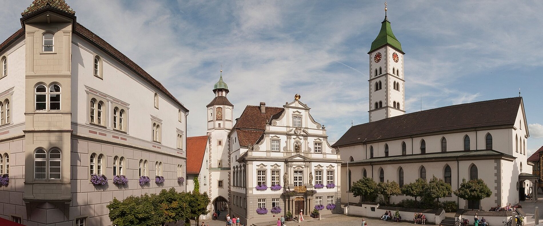 Panoramaansicht eines Platzes mit historischen Gebäuden, darunter ein Rathaus und eine Kirche mit markantem Turm. Die Architektur zeigt eine Mischung aus traditionellem und modernem Stil, umgeben von Bäumen und Blumen. Wolken am Himmel tragen zur Atmosphäre des Tages bei.