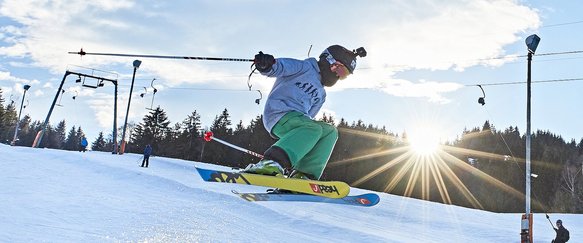 Ein Skifahrer springt über den Schnee, während die Sonne im Hintergrund aufgeht. Die Szene zeigt die Bewegungsdynamik und einen klaren Himmel mit einigen Wolken. Der Skifahrer trägt eine graue Jacke, grüne Hose und eine Mütze.