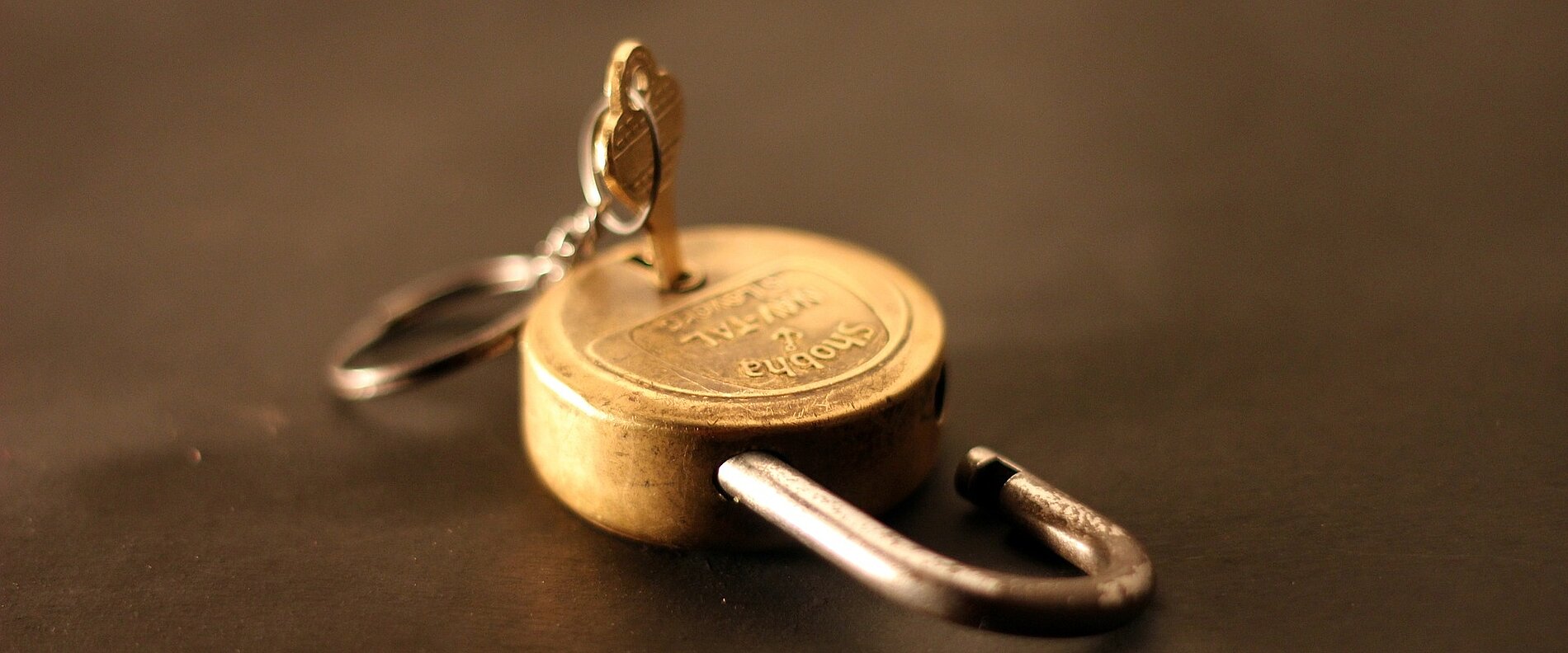Ein offenes Vorhängeschloss mit einem Schlüssel, der steckt. Der Schlüssel hängt an einem kleinen Ring. Der Hintergrund ist dunkel, was den Fokus auf das Schloss und den Schlüssel legt.