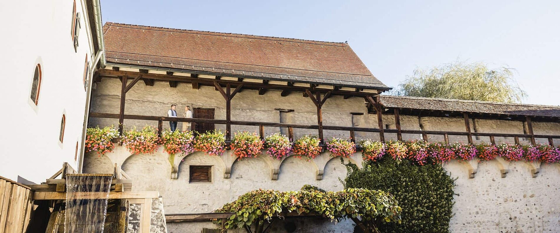 Eine historische Mauer mit einer Holzveranda, die mit bunten Blumen dekoriert ist. Auf der Veranda steht eine Person. Im Hintergrund sind Bäume und ein blauer Himmel sichtbar.