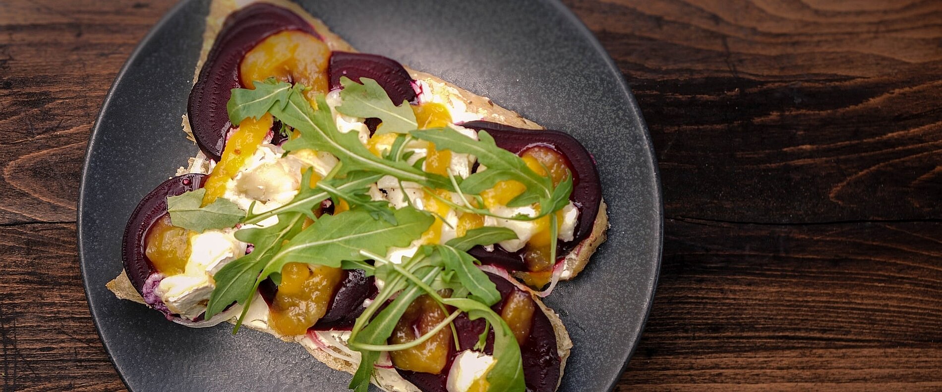 Ein Teller mit einer belegten Brotscheibe, belegt mit roten Zwiebeln, Orangenscheiben und einem frischen Rucola-Servier. Die Kombination wirkt appetitlich und farbenfroh auf einem dunklen Holzuntergrund.