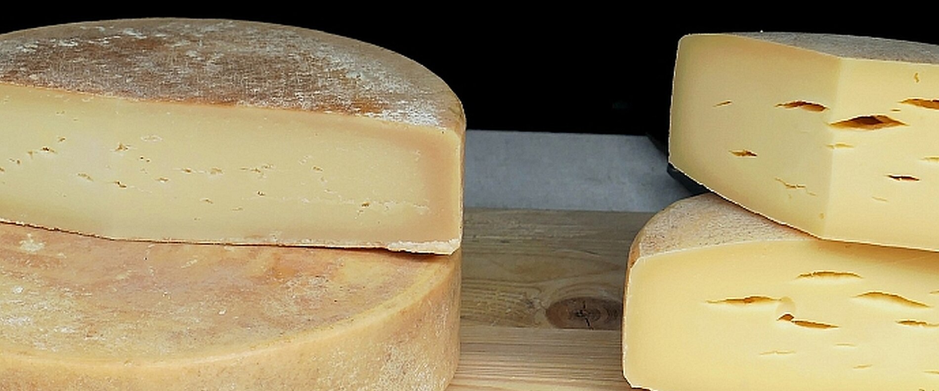 Zwei große Käselaibe liegen auf einem Holzbrett. Einer der Käse ist ganz, der andere ist in Stücke geschnitten. Die Käsesorten zeigen eine goldene Farbe und eine glatte Oberfläche mit kleinen Löchern.