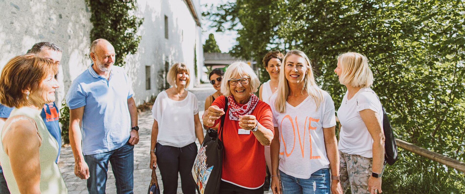 Eine Gruppe von zehn Personen steht in einem grünen Außenbereich. Einige Lächeln und unterhalten sich, während eine Dame mit Brille und rotem Oberteil eine Tasse hält. Im Hintergrund ist ein helles Gebäude und Bäume zu sehen. Die Atmosphäre wirkt freundlich und entspannt.
